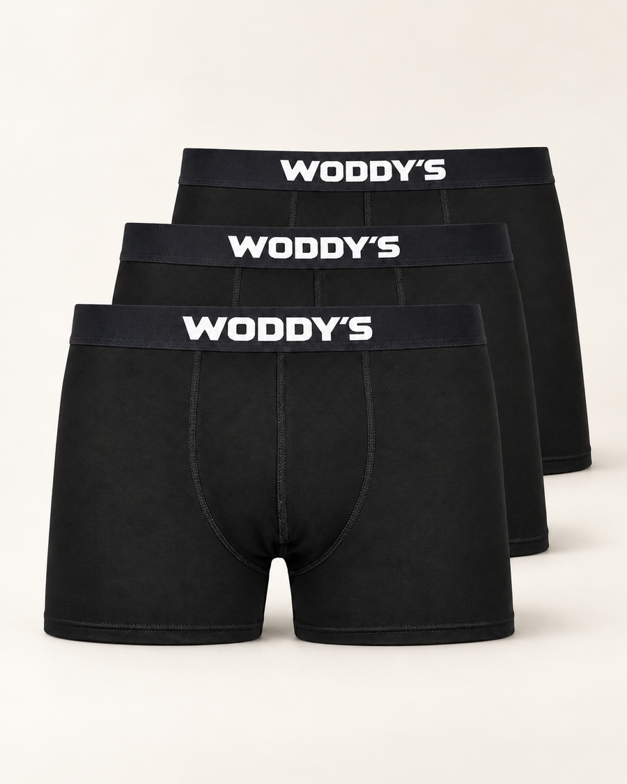 Drei schwarze Boxershorts mit kurzem Bein und weißem WODDY’S-Logo, versetzt vor warmem Off-White.